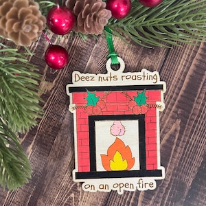 May include: Wooden Christmas ornament shaped like a fireplace with the text "Deez nuts roasting on an open fire." The fireplace is red brick with a black frame, a fire, and a humorous design.