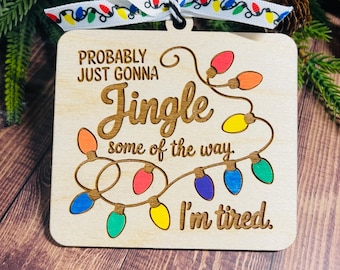 Puede incluir: Adorno navideño de madera con el texto "Probably just gonna Jingle some of the way. I'm tired." El adorno presenta una cadena de luces navideñas de colores y una cinta decorativa.