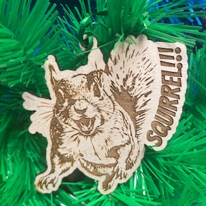 May include: A wooden Christmas ornament featuring a squirrel with a toothy grin and the word "SQUIRREL!!" written on the side. The ornament is hanging on a green artificial Christmas tree.