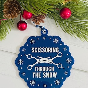 May include: A blue Christmas ornament with white text that reads "SCISSORING THROUGH THE SNOW" and a pair of scissors. The ornament is decorated with snowflakes and hangs from a white and blue string. The background includes pine needles, pine cones, and red ornaments.