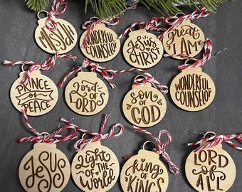 Op de afbeelding: Houten kerstversieringen met gegraveerde teksten zoals "Jezus Christus" en "Koning der Koningen". Elke ronde ornament is voorzien van rood-wit touw om op te hangen. De ornamenten zijn gemaakt van lichtkleurig hout.