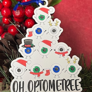 May include: A wooden Christmas ornament shaped like a tree, featuring cartoon eyeballs with festive accessories. The ornament has the text "OH OPTOMETREE" in black. The eyes have Santa hats, scarves, and reindeer antlers.