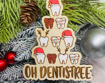 Puede incluir: Adorno navideño de madera con forma de árbol de Navidad, con dientes con gorros de Papá Noel, cuernos de reno y luces de cadena. El texto "OH DENTISTREE" está grabado en la madera. Se adjunta una cinta roja para colgarlo.