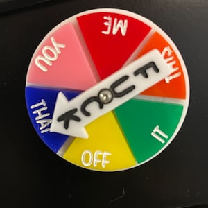 Puede incluir: Un juego circular colorido con una flecha blanca y la palabra "FUCK" en él. El juego está dividido en segmentos de rojo, naranja, verde, amarillo, rosa y azul, cada uno con una palabra. Las palabras son "ME", "THIS", "IT", "OFF", "YOU" y "THAT".