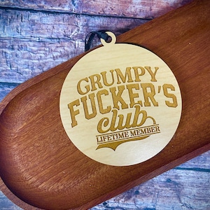 May include: A circular light-colored wooden ornament featuring the engraved text "GRUMPY FUCKER'S club" in a bold, stylized font, with the words "LIFETIME MEMBER" printed below, and a small black ribbon loop at the top for hanging.
