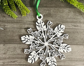 May include: White snowflake ornament with a green ribbon for hanging. The ornament features a dinosaur skeleton design surrounded by leaf shapes. The ornament is made of a light-colored material.