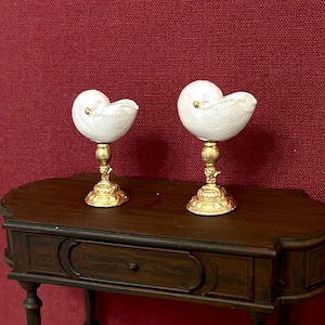 Op de afbeelding: Een miniatuur donkerbruine houten sidetable met één lade en vier slanke poten, voorzien van een onderste plank. Twee witte nautilus-schelpvormige decoratieve objecten met goudkleurige voetstukken staan op het tafelblad.