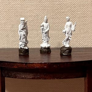 Op de afbeelding: Drie kleine, witte beelden van Aziatische figuren staan op een donkere houten tafel. Elk beeld is ingewikkeld gedetailleerd en staat op een donkerbruine basis. De beelden zijn van verschillende figuren, elk met unieke poses en kleding.