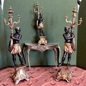 Könnte beinhalten: Miniatur-Kerzenständer und Figuren im antiken Stil. Die schwarzen Figuren halten verzierte goldene Kerzenständer. Die Figuren tragen goldene Röcke. In der Mitte steht ein kleiner Tisch mit Marmorplatte und goldenen Verzierungen. Der Hintergrund ist grün.