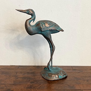 Puede incluir: Estatua de bronce de una garza con acabado de pátina. El ave se representa en una pose elegante, de pie sobre una pata con la cabeza y el pico extendidos. La estatua tiene un aspecto envejecido, lo que aumenta su encanto antiguo. Un acento decorativo para el hogar o el jardín.