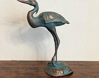 Puede incluir: Estatua de bronce de una garza con acabado de pátina. El ave se representa en una pose elegante, de pie sobre una pata con la cabeza y el pico extendidos. La estatua tiene un aspecto envejecido, lo que aumenta su encanto antiguo. Un acento decorativo para el hogar o el jardín.