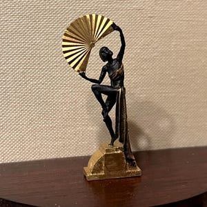 Peut inclure: Figurine Art Déco noire et dorée représentant une danseuse avec un éventail. La figurine est posée sur un socle doré, avec un éventail doré tenu en l'air. La robe et le socle de la danseuse ont des détails dorés.