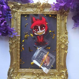 May include: A gold-colored ornate picture frame with a cartoon character keychain and a small plastic container with a fork. The keychain features a red character with a sinister grin. The frame has a dark gray background with gold confetti.