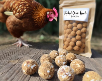 May include: A brown hen with a red comb and wattle stands on a wooden surface. In front of the hen is a bag of rolled chick treats labeled "Rolled Chick Treats, Beer & Seeds". The bag is open and the treats are visible. The treats are small, round, and brown with white specks.