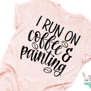 Op de afbeelding: Lichtroze t-shirt met de zwarte tekst "I RUN ON coffee & painting". Koffieboonafbeeldingen zijn door de tekst heen verwerkt. Het shirt heeft opgerolde mouwen en een ronde hals.