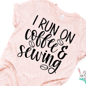 Op de afbeelding: Lichtroze t-shirt met de zwarte tekst "I RUN ON coffee & sewing". Koffieboongraphics zijn inbegrepen. Het shirt heeft korte mouwen en een ronde hals.