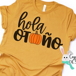 Op de afbeelding: Een mosterdgeel T-shirt met de tekst "hola otoño" in zwarte letters en een kleine oranje pompoenafbeelding. De tekst is in een cursief schrift.