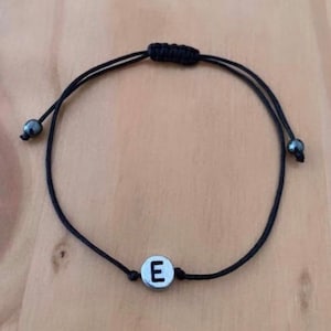 May include: A black string bracelet with a silver charm featuring the letter 'E'.