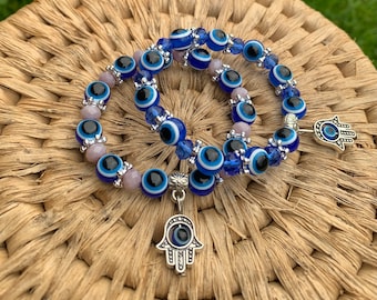 Peut inclure: Deux bracelets bleu œil-de-dieu avec des breloques hamsa argentées. Les bracelets sont faits de perles de verre bleues et blanches et de perles argentées. Les breloques hamsa sont argentées et ont un œil-de-dieu bleu au centre.