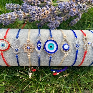 May include: A set of ten colourful bracelets with different charms, including evil eye, hamsa hand, and nautical wheel designs. The bracelets are made with blue and red string.