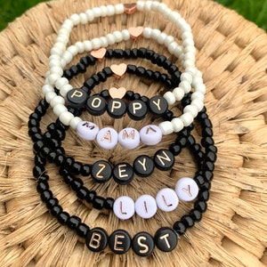 May include: Five beaded bracelets with black and white beads and gold lettering spelling out the names "Poppy", "Mama", "Zeyn", "Lily", and "Best".