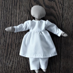 May include: A white fabric doll with a brown head and long sleeves. The doll is wearing a white dress and pants.
