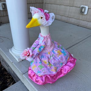 May include: A decorative goose figurine wearing a pink dress with butterfly patterns and a matching bonnet. The goose holds a pink rose and has a yellow beak. The dress has a satin trim.