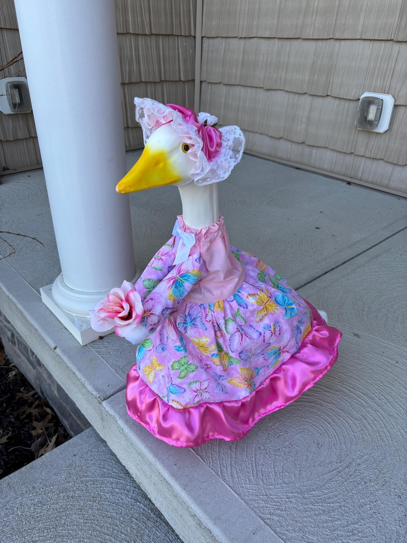 May include: A decorative goose figurine wearing a pink dress with butterfly patterns and a matching bonnet. The goose holds a pink rose and has a yellow beak. The dress has a satin trim.