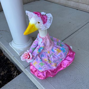 May include: A decorative goose figurine wearing a pink dress with butterfly patterns and a matching bonnet with lace trim. The goose holds a pink rose and has a yellow beak.