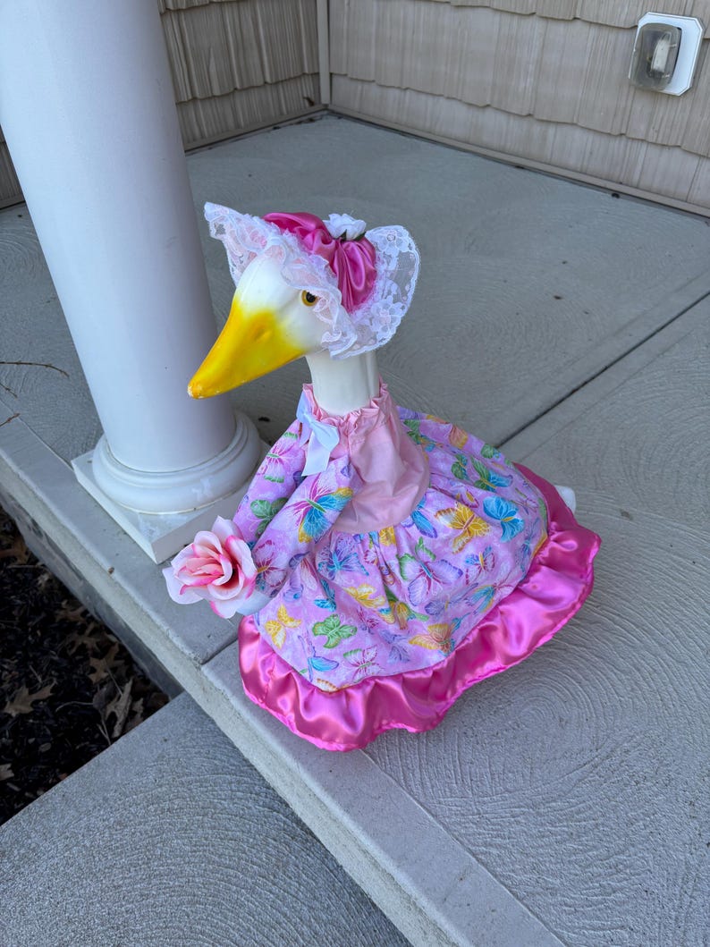 May include: A decorative goose figurine wearing a pink dress with butterfly patterns and a matching bonnet with lace trim. The goose holds a pink rose and has a yellow beak.