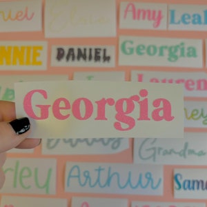 May include: A collection of white stickers with various names written in different colours and fonts. The names include Ashley, Mummy, Ralph, Amy Leah, Daniel, ONNIE, PANIEL, Georgia, E, Georgia, Ley, Arthur, Bonnie, Amy, ISOBEL, WILLIAMS, James, and FRID.