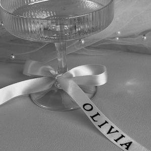 May include: A clear glass champagne flute with a white satin ribbon tied around the stem. The ribbon has the name "Olivia" printed on it in black.