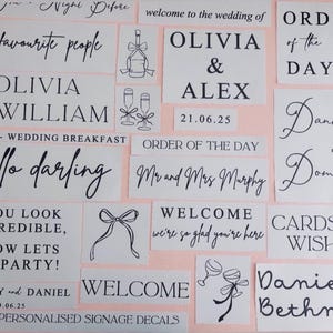 May include: A selection of white rectangular decals with black text and illustrations, displayed on a pale pink background. The decals include wedding-themed phrases, names, dates, and decorative elements like champagne flutes and bows.