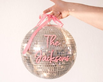 Könnte beinhalten: Eine kugelförmige Discokugel, die mit kleinen reflektierenden silbernen Spiegelkacheln bedeckt ist. Die Kugel hat ein oben gebundenes rosa Band und eine rosa Schrift auf der Vorderseite, die The Jacksons lautet.