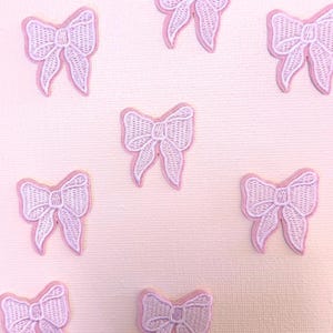 May include: Embroidered patches in the shape of bows, in light pink. Each patch has a detailed bow design with a white centre and a pink outline. The patches are scattered on a light pink background.
