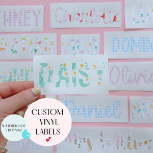 May include: A selection of custom vinyl labels featuring names in diverse fonts and patterns. The label held displays "DAISY" in mint green with white daisy accents. The labels are waterproof and durable, suitable for various applications.