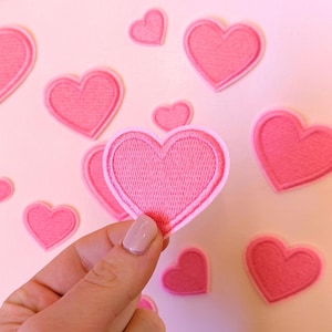 May include: A collection of pink embroidered heart patches of varying sizes, scattered on a pale pink background. One patch is held by a hand. Each patch features a stitched outline and a filled-in centre, suitable for crafting and DIY projects.