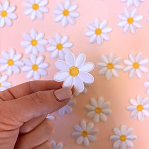 May include: Embroidered white daisy flower patches with yellow centres. The image shows a hand holding one patch, with several more scattered on a pink surface. These patches are ideal for adding a floral touch to clothing or accessories. Each patch has a detailed design.