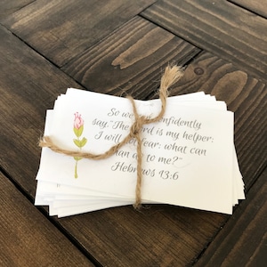 A stack of 40 printable encouraging cards with bible verses is tied together with twine. The cards sit on a dark brown wooden table top. The design on the card is a beautiful watercolor flower with a handwritten script.