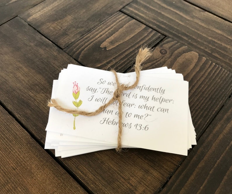 A stack of 40 printable encouraging cards with bible verses is tied together with twine. The cards sit on a dark brown wooden table top. The design on the card is a beautiful watercolor flower with a handwritten script.