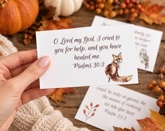 Könnte beinhalten: Eine rechteckige weiße Karte mit einer Aquarellillustration eines Fuchses und dem Text: O Lord my God, I cried to you for help, and you have healed me. Psalms 30:2. Die Karte wird vor einem Hintergrund aus Herbstblättern und Kürbissen gehalten.