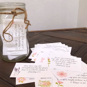 Encouraging scripture cards with watercolor flower designs and handwritten font lay across a dark wood table. A glass mason jar with twine tied around the top is filled with the rest of the scripture cards from the set.