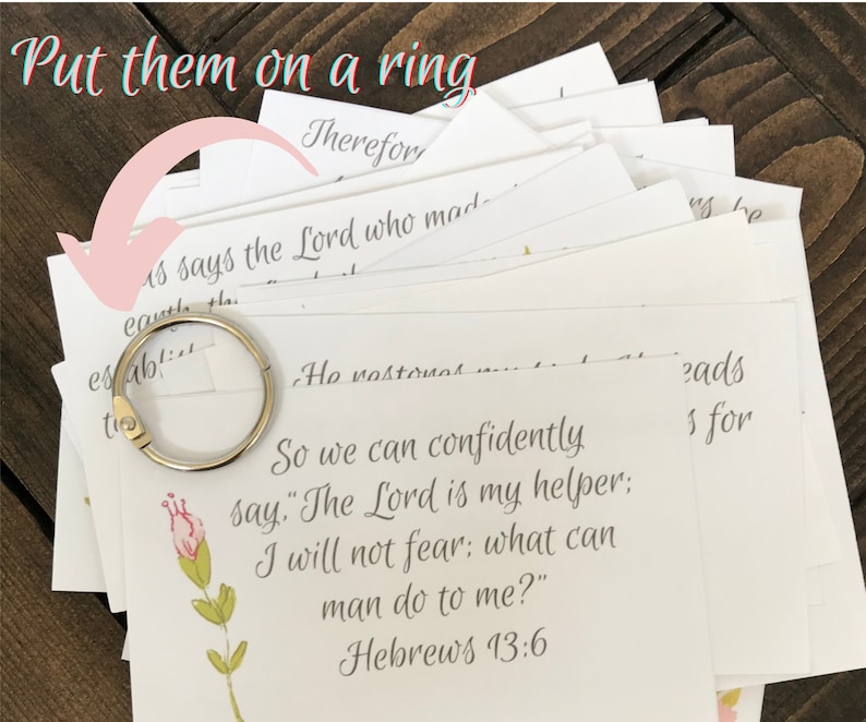 Stack of small bible verse cards sitting on a dark brown wood table. The first card has a pink flower that is on a green stem. a silver book ring lies on the top of the stack of cards. The notes on the photo says "put them on a ring"
