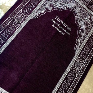 Op de afbeelding: Een dieppaars gebedskleed met een decoratieve witte rand en een centraal boogontwerp. Het kleed heeft de tekst "Hawarun May all your duas be accepted" in witte letters. Het kleed is rechthoekig.
