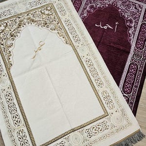 Op de afbeelding: Twee gebedskleden, &eacute;&eacute;n cr&egrave;mekleurig en &eacute;&eacute;n dieppaars, worden getoond. Het cr&egrave;mekleurige kleed heeft ingewikkelde bruine en gouden patronen en Arabisch schrift. Het paarse kleed heeft witte patronen en Arabisch schrift. Beide hebben gefranjerde randen.