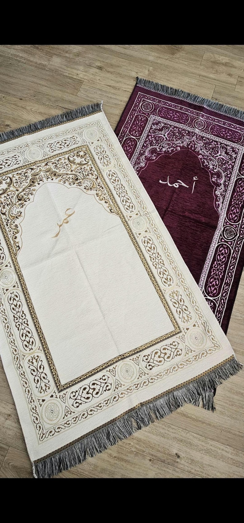 Op de afbeelding: Twee gebedskleden, &eacute;&eacute;n cr&egrave;mekleurig en &eacute;&eacute;n dieppaars, worden getoond. Het cr&egrave;mekleurige kleed heeft ingewikkelde bruine en gouden patronen en Arabisch schrift. Het paarse kleed heeft witte patronen en Arabisch schrift. Beide hebben gefranjerde randen.