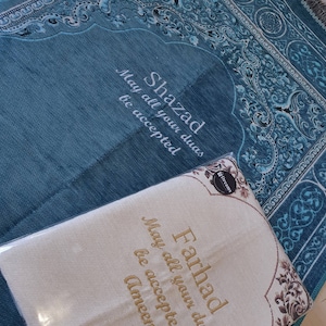 Op de afbeelding: Een turquoise gebedskleed met een decoratieve rand en de naam "Shazad" in het wit geborduurd, samen met de zin "May all your duas be accepted." Een cr&egrave;mekleurig gebedskleed met de naam "Farhad" en dezelfde zin is ook zichtbaar.