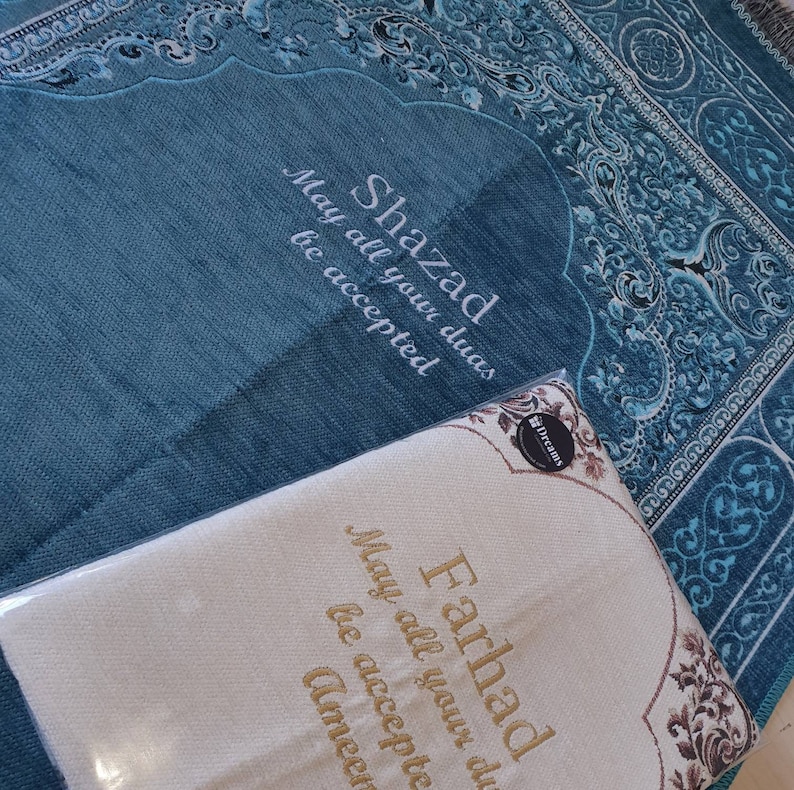 Op de afbeelding: Een turquoise gebedskleed met een decoratieve rand en de naam "Shazad" in het wit geborduurd, samen met de zin "May all your duas be accepted." Een cr&egrave;mekleurig gebedskleed met de naam "Farhad" en dezelfde zin is ook zichtbaar.