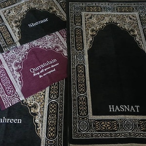 Op de afbeelding: Verschillende gebedskleden in zwart, bordeaux en grijs met sierlijke randontwerpen. De kleden hebben boogvormige ontwerpen en de namen "Shezaar", "Qurratulain", "Mahreen" en "Hasnat" in witte letters. Het bordeauxkleurige kleed heeft de tekst "May all your duas be accepted".