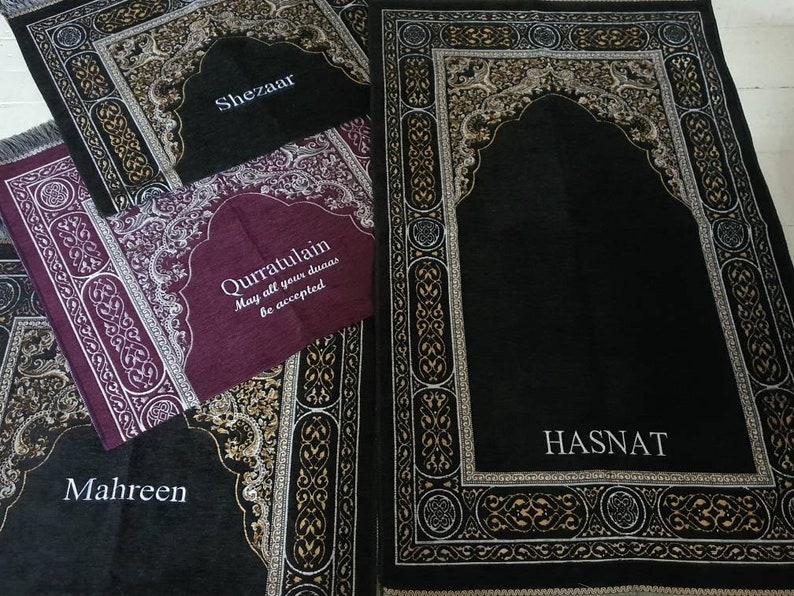 Op de afbeelding: Verschillende gebedskleden in zwart, bordeaux en grijs met sierlijke randontwerpen. De kleden hebben boogvormige ontwerpen en de namen "Shezaar", "Qurratulain", "Mahreen" en "Hasnat" in witte letters. Het bordeauxkleurige kleed heeft de tekst "May all your duas be accepted".